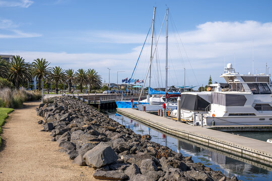 Wyndham Harbour marina in Werribee South, Victoria, Australia, moored sailing yachts along a concrete pontoon jetty. A rocky stone seawall lines the foreshore pathway. Coastal leisure infrastructure