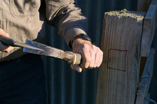 Fence workman using a hammer and chisel to work on a wooden gate post on a farm