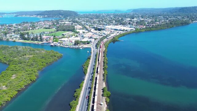 Drone aerial pan of a freight train carrying shipping containers on tracks entering Woy Woy surrounded by urban town buildings and Brisbane Water waterfront foreshore on the Central Coast Australia