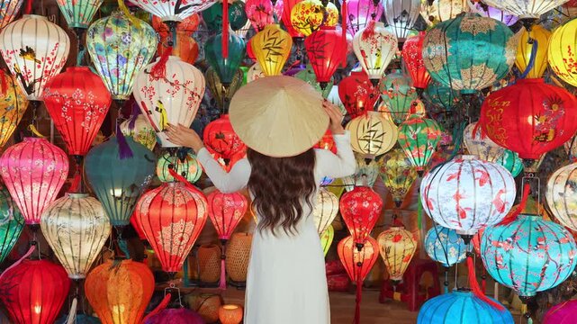 Slow-motion video of a young woman in a traditional white Vietnamese dress, wearing a conical hat, amidst a display of colorful lanterns in the ancient town of Hoi An, Vietnam.