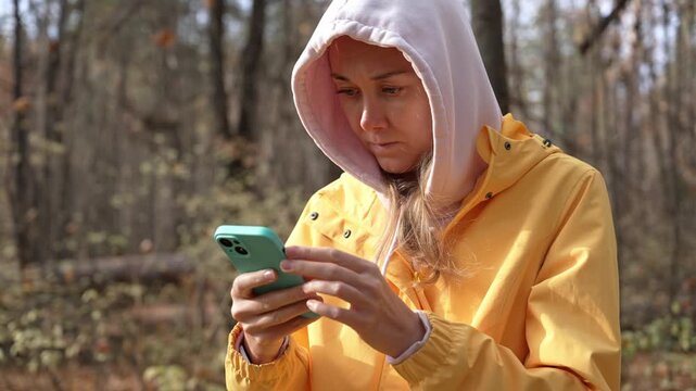 Young woman in a yellow raincoat and hoodie standing in an autumn forest while using her mobile phone for navigation, looking at the map with a concentrated expression on her face