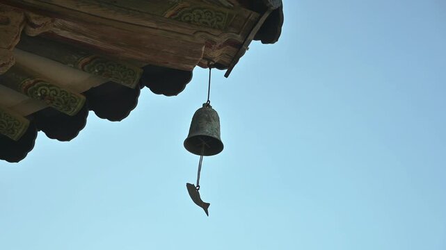 푸른 하늘을 배경으로 법주사 팔상전 목조 처마 밑에서 흔들리는 물고기 풍경.(Wind chime with fish pendant swaying under Beopjusa Palsangjeon wooden eaves against blue sky.)