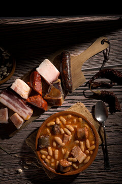 Traditional steaming hot Spanish fabada asturiana stew in clay bowl with pork meats chorizo and blood sausage on rustic wooden table