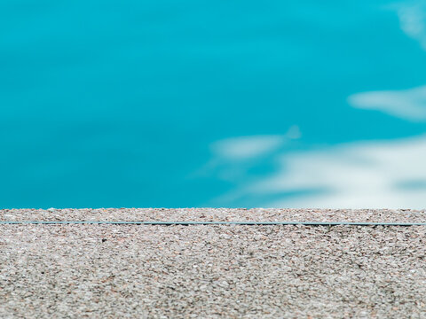 Close-up of textured pebble concrete pool edge beside clear turquoise water with soft cloud reflections, minimalist summer swimming pool background