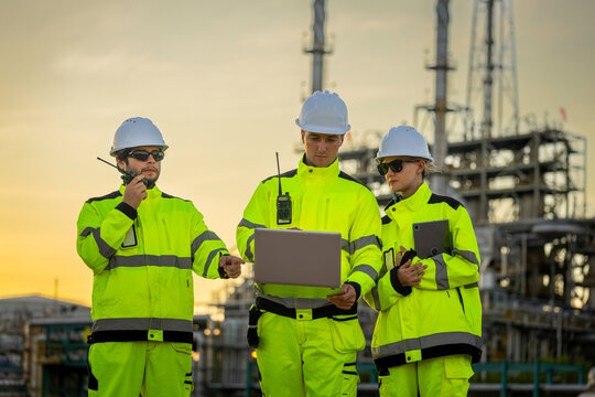 Industrial engineers using laptop, tablet and walkie talkie at refinery plant at sunset, Engineering team in safety high-visibility jackets inspecting chemical refinery at dusk