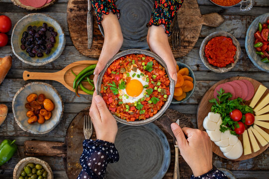 Shakshuka, fried eggs with tomatoes, onion, red pepper and spices in cast iron pan, other name is menemen. Hands of two women sharing and holding their meal.  