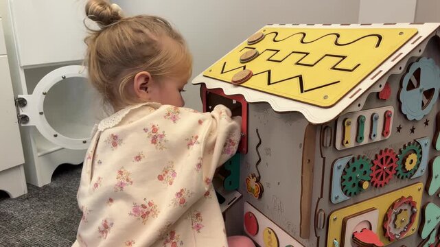 A girl opens a flap on a wooden activity house and reaches toward roof sliders, sensory play, childhood learning, motor skills.