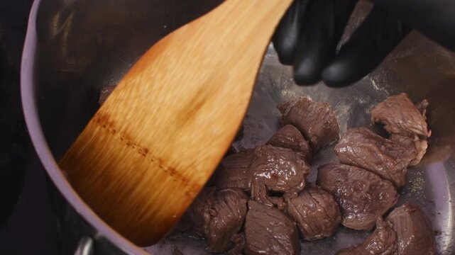 A black-gloved hand stirs browned beef chunks in a steel pan using a wooden spatula during cooking.