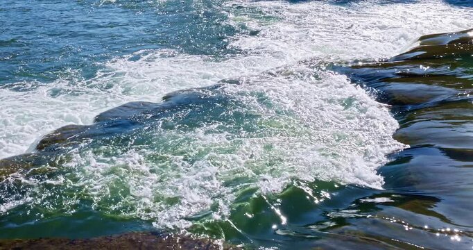 Rush water flowing rapidly over dark rocks creating white foam in bright sunlight.