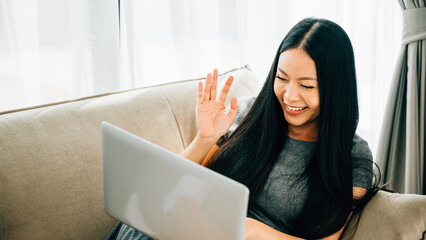 Asian woman on sofa uses laptop for video conferencing greeting colleague © sorapop