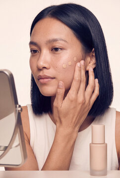 Woman applying foundation drops to her face in a skincare routine with bottle beside mirror for beauty banner and cosmetic product layout