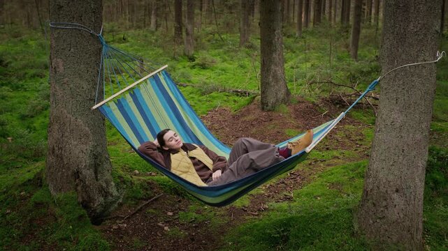 Caucasian young adult woman resting and closing eyes in hammock hanging between trees in mossy forest. Footage fits mental wellbeing, stress relief and ecotourism campaigns