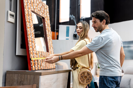 Young couple measuring a decorative mirror with a tape measure, selecting new items for their home during a shopping trip