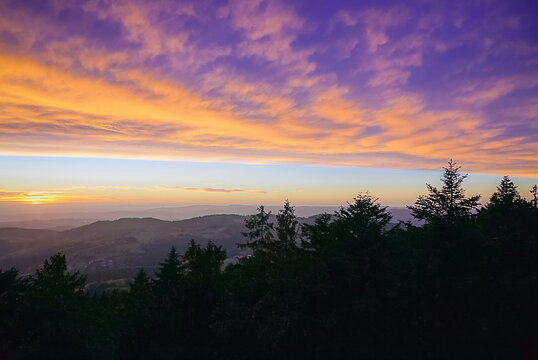 Dramatic autumn sunset over the Bavarian Forest in Germany with colorful purple and orange clouds, distant mountain ridges and dark evergreen trees.