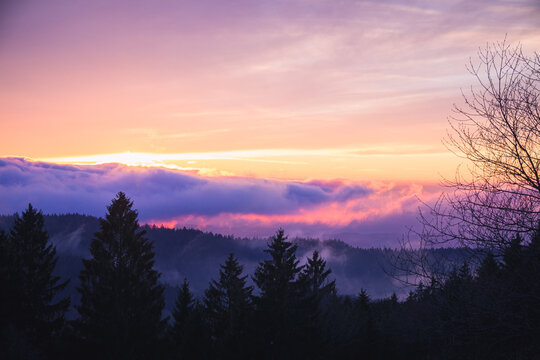 Glowing orange sunset above the Bavarian Forest in Germany with dramatic cloud formations, rolling mountain scenery and evergreen trees in the foreground.