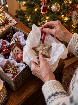 Wrinkled hands unwrapping a worn wooden star ornament from tissue beside a Christmas tree and a storage box of decorations, showing gentle care for a keepsake in a warm home with soft lights