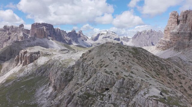 Aerial drone view gliding over a broad grey limestone ridge toward a lone blocky rock tower with wide high alpine plateaus and distant peaks beyond near Tre Cime di Lavaredo in South Tyrol Italy