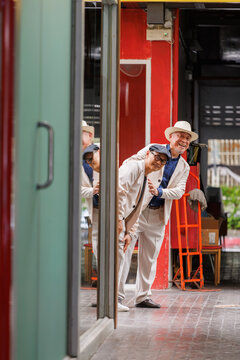 Two senior gay men lean from behind doorway with cheerful expressions, moving through colorful shopfronts, narrow walkways together whilst making travel memories through time spent together.