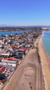 Long Beach, California, USA - October 15, 2026: Vertical view of dump trucks hauling sand along a flood berm between Peninsula homes and ocean, Alamitos Bay behind, after Tropical Storm Rachel.