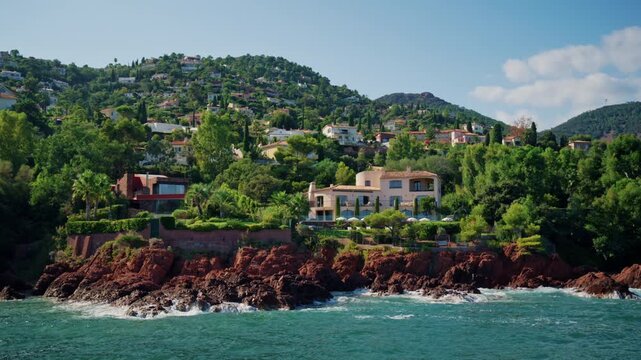 Pink seaside villa with gardens and parasols above red rocks and crashing waves, with houses on green hills behind, Esterel, France.