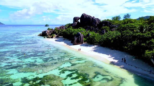 Bright daytime brings clear turquoise water, a shallow coral reef and pale sand to La Digue Island, Seychelles, where visitors stroll beneath lush greenery and dramatic granite boulders.