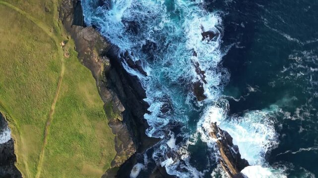 Orkney Mainland - Scotland- Aerial view of rugged coastal cliffs with crashing waves. Green grass covers the cliffside, contrasting with dark rock. Ocean water churns with white foam as it hits the sh