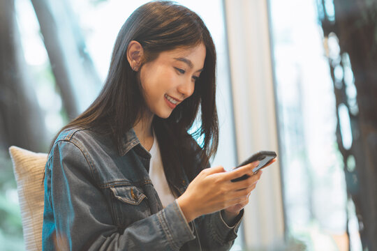 Smiling young Asian woman using smartphone with both hands while sitting indoors near bright window, enjoying mobile communication, online connectivity