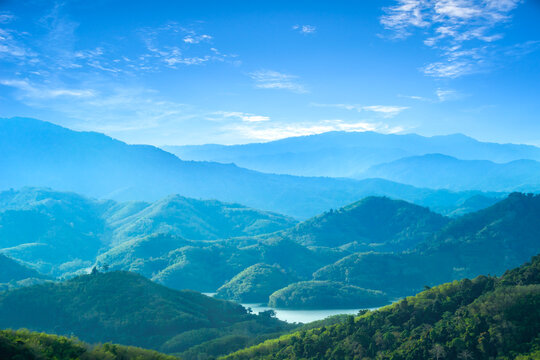 A sunny landscape featuring lush green vineyards, forests, and farmhouses under a blue sky.
