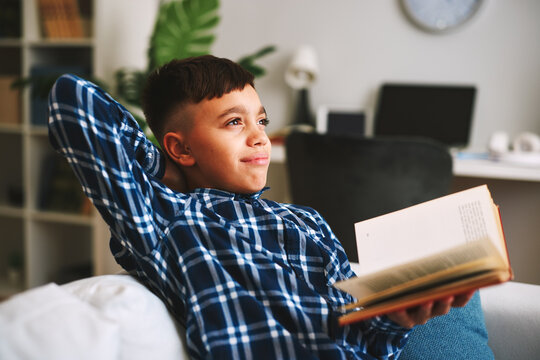 Young boy relaxing on sofa with open book, gazing upward and daydreaming about the future, finding inspiration and joy in reading and learning at home