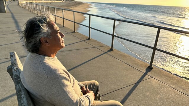 Sitting senior Asian man wearing beige sweater and pants on promenade bench by railing, copy space
