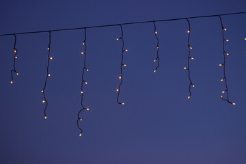 Golden icicle fairy lights hang against a clear blue twilight sky creating a festive outdoor decoration with warm glowing bulbs and generous copy space for seasonal greetings and event designs © Aleksey Konovaloff