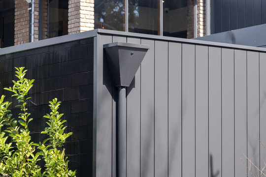 A sleek dark metal rain hopper head connected to a round downpipe for stormwater drainage on exterior wall of a residential house in Australia.