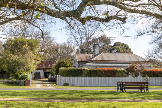 A heritage weatherboard cottage with an aged corrugated iron roof and brick chimneys sits behind a white timber picket fence in a quiet Australian country town. Well-preserved historic character 
