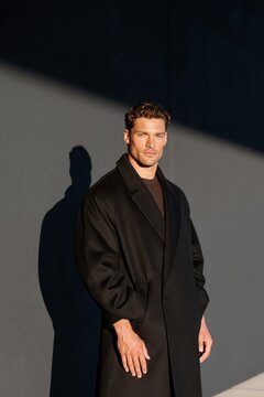 Man in long black coat standing against gray wall with dramatic side lighting casting sharp shadow, confident expression, minimalist urban style