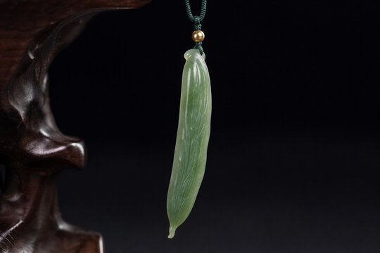 A slender green jade pendant shaped like a silique on a dark background.