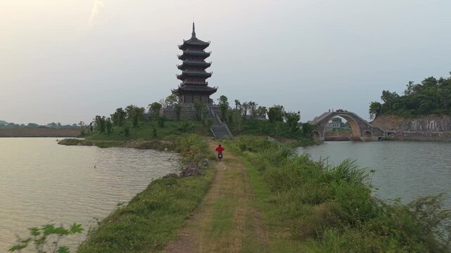 Lone cyclist riding a bicycle along a narrow dirt path towards the historic pagoda zoe mascot, surrounded by tranquil water in vietnam