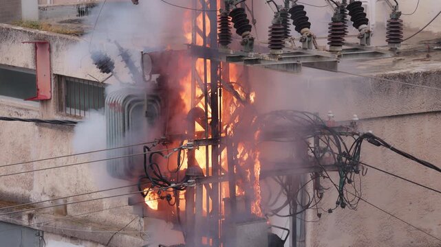 Video shot of a high voltage power pole and electrical transformer burning with intense flames, sparks, and thick white smoke on a city street after a short circuit