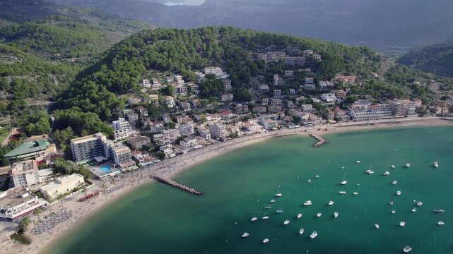Drone shot showcases Soller&rsquo;s scenic coastline, marina with anchored boats, lush hills, and Mediterranean resort buildings under bright daylight in stunning landscape composition.