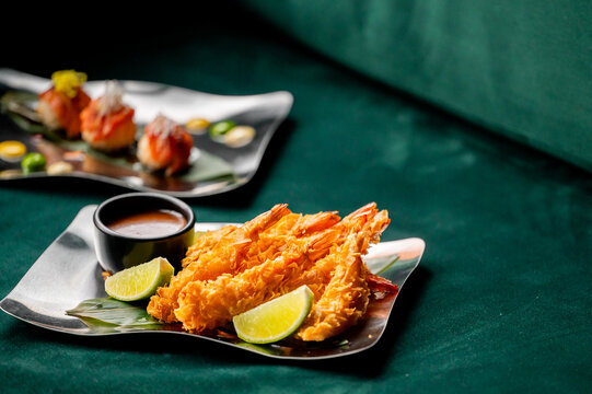 Crispy fried ebi tempura shrimp served on a metal plate with lime slices and dipping sauce on a dark green fabric background.