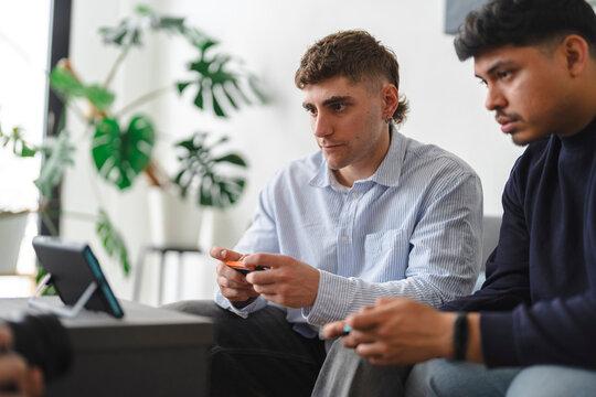 Two young men sit together in a bright living room, playing a video game on a portable console. They hold controllers and watch the small screen with concentrated expressions.