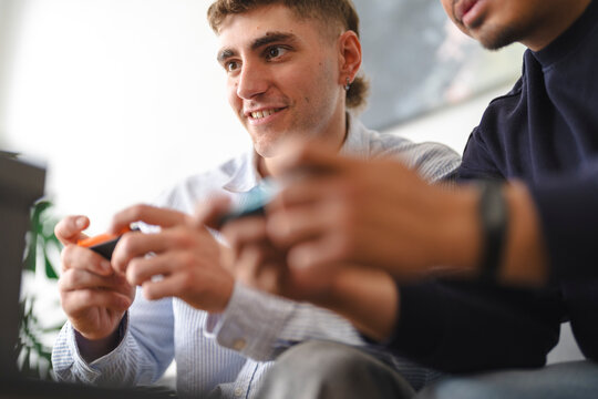 Two young male friends are sitting together on a couch indoors, holding handheld controllers and smiling while playing video games during their leisure time.