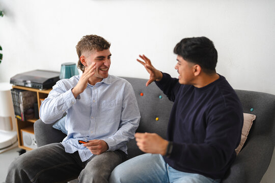 Two young men are sitting together on a grey sofa in a bright living room, laughing and gesturing playfully while having a fun conversation during their indoor hangout.