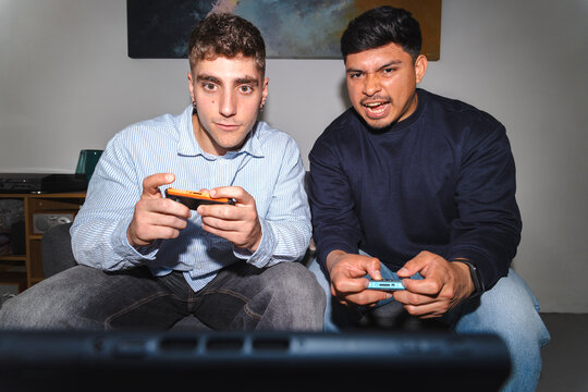 Two young male friends sitting on a couch in a living room, holding video game controllers. They are playing a game together with intense and excited expressions on their faces.