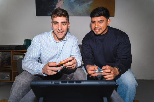Two young male friends sitting on a living room couch together. They are smiling while playing a video game on a portable console device, enjoying free time at home.