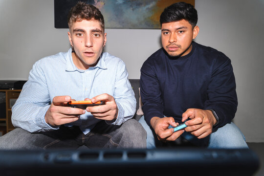 Two young men sitting on a couch in a living room, playing a video game together. They hold handheld controllers and watch the screen with focused and surprised expressions.