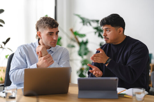 Two male coworkers sit together at an office desk, collaborating on a project while looking at a computer. They converse and gesture actively during a business meeting.