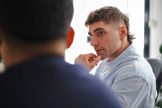 Two young male colleagues having a serious discussion in a bright modern office. One man listens intently with his hand on his chin while facing his coworker during a meeting.