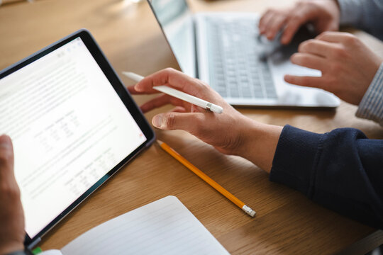 Two professionals collaborate in an office workspace. One person reviews data on a digital tablet using a stylus while a colleague types on a laptop nearby.