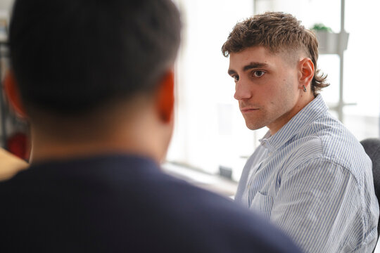 Two young men are sitting together in a bright office. One man in a striped shirt listens intently while facing his coworker during a professional conversation.