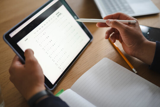 A person sits at a wooden desk, holding a digital tablet with a stylus. They review data displayed on the screen while an open notebook and a laptop rest nearby.